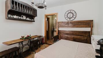 A bedroom with a wooden bed, a wooden desk, a shelf with decorative items, a wall clock, and a door leading to another room.