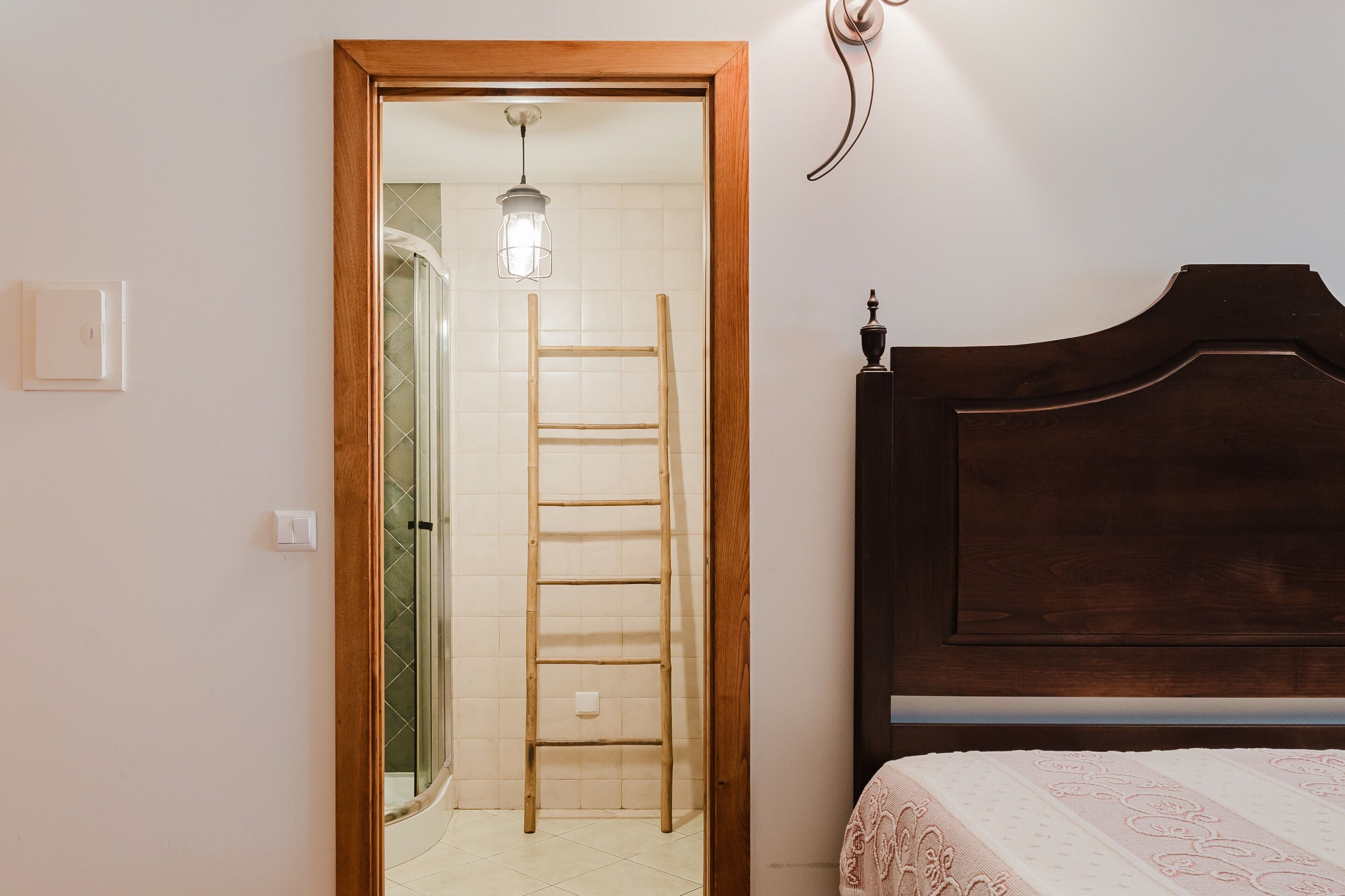 A bedroom with a wooden bed frame, a ladder in the doorway, and a light fixture on the wall.