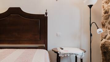 A room with a wooden bed, a small table with a white tablecloth, a lamp, and a stone wall.