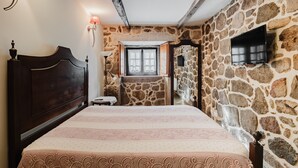 A bedroom with a stone wall, a bed with a patterned bedspread, a wooden headboard, a bedside table, a lamp, and a TV mounted on the wall. - Room 'Duplo - 4' with Shared Terrace and Wi-Fi (Pitões das Junias)