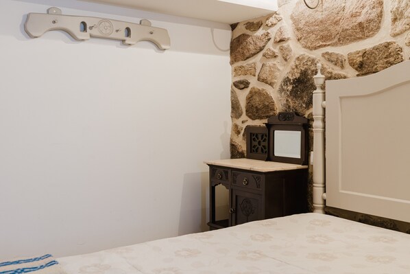Een slaapkamer met een stenen muur, een bed met een bloemenbedekking, een dressoir met spiegel en een wandplank.