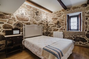 A bedroom with a stone wall, a bed with a white headboard, a nightstand, a dresser, and a window with a view.