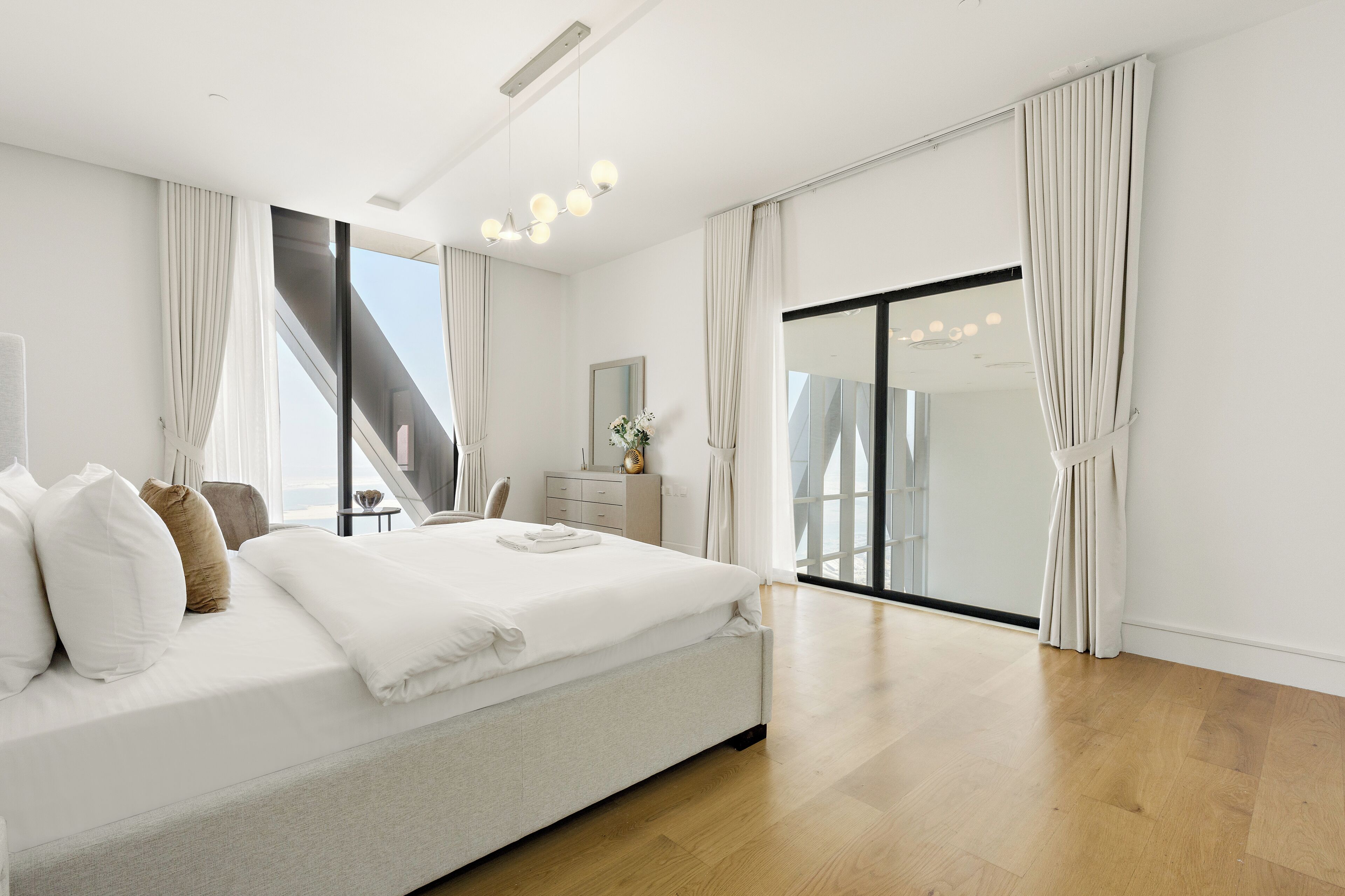 A modern bedroom with a large bed, a dresser, a mirror, and a sliding glass door leading to a balcony.