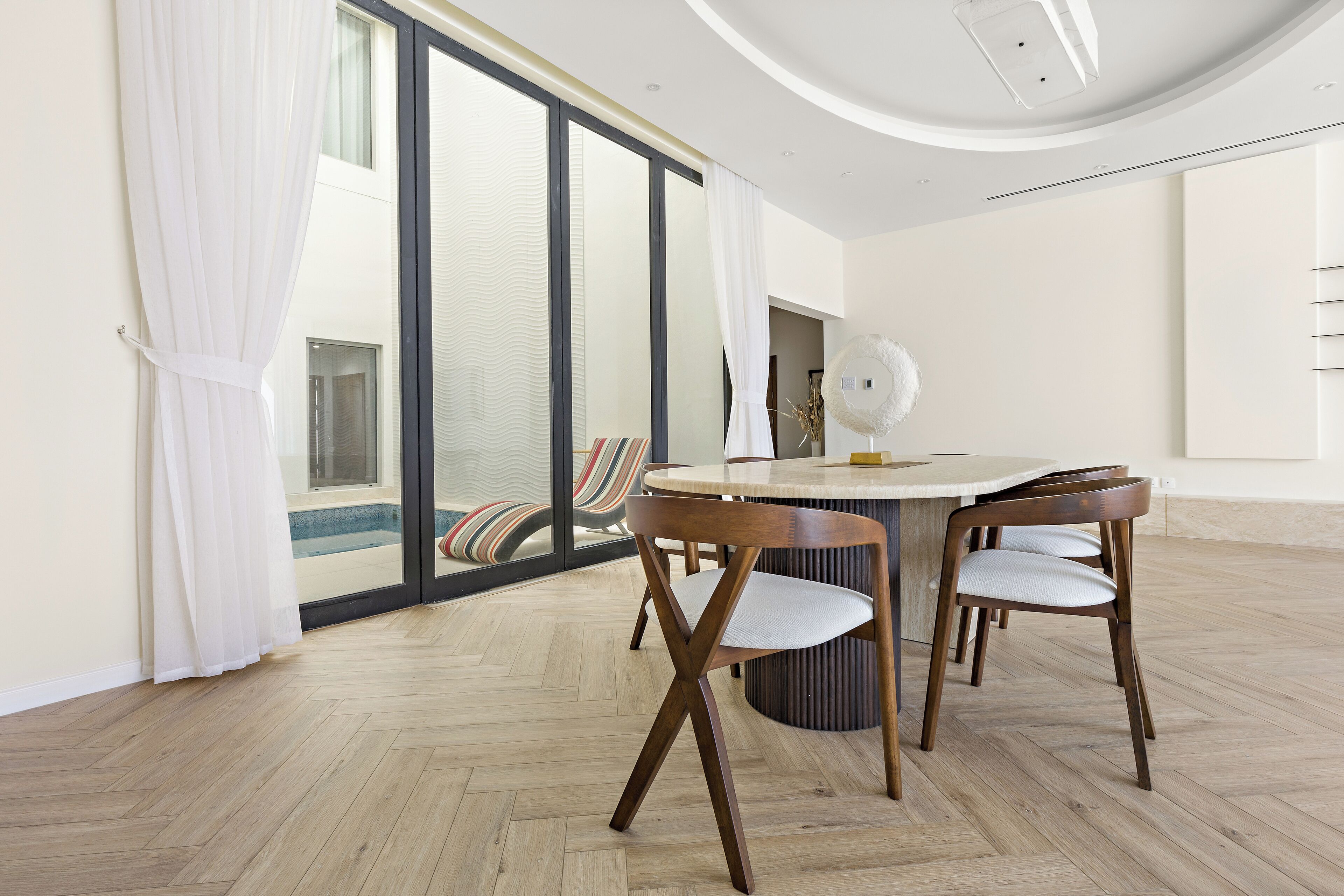 Un coin repas avec une table ronde et quatre chaises en bois, de grandes fenêtres avec des rideaux et une vue sur une piscine.
