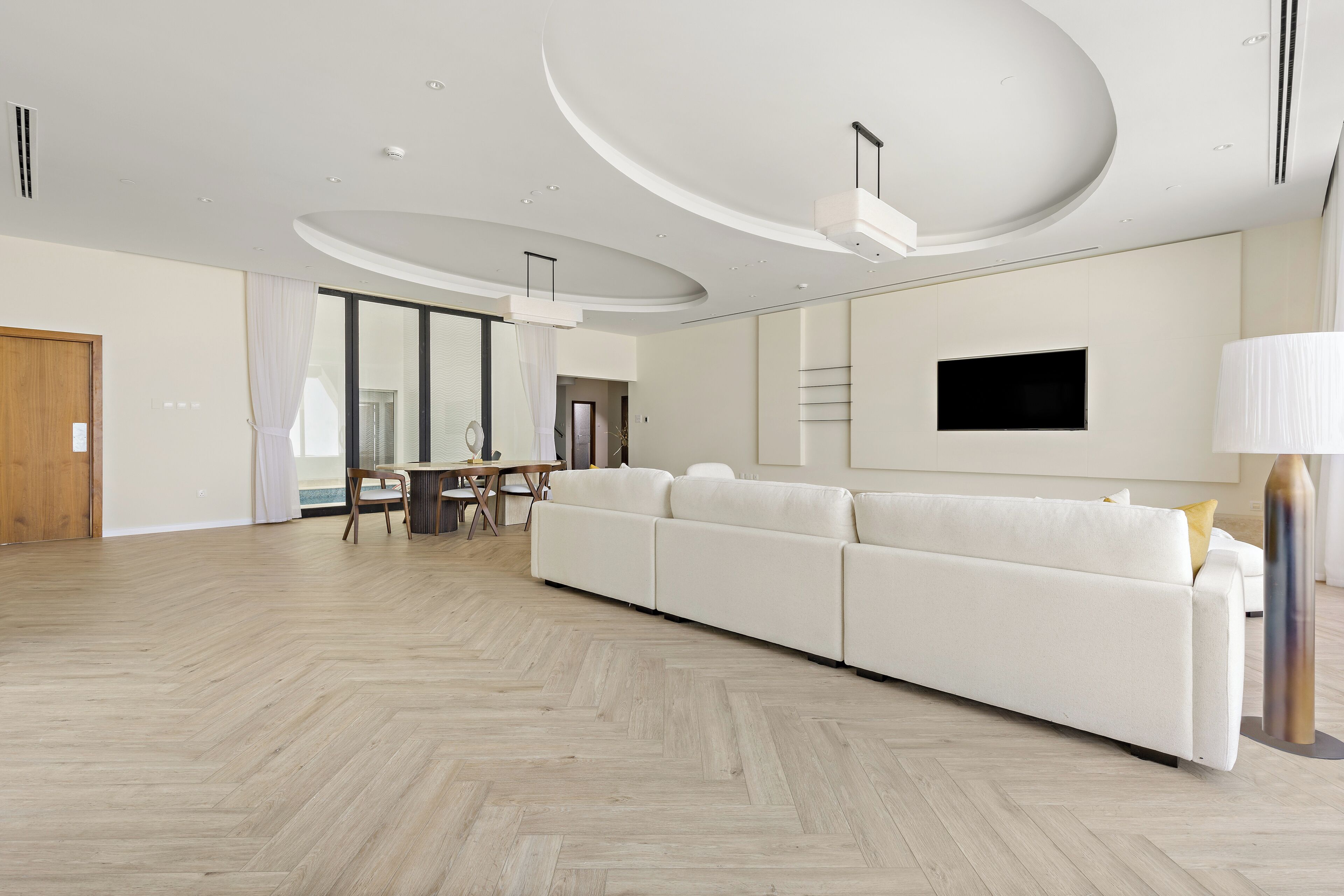 A spacious living room with a large white sofa, a flat-screen TV mounted on the wall, and a herringbone-patterned wooden floor.