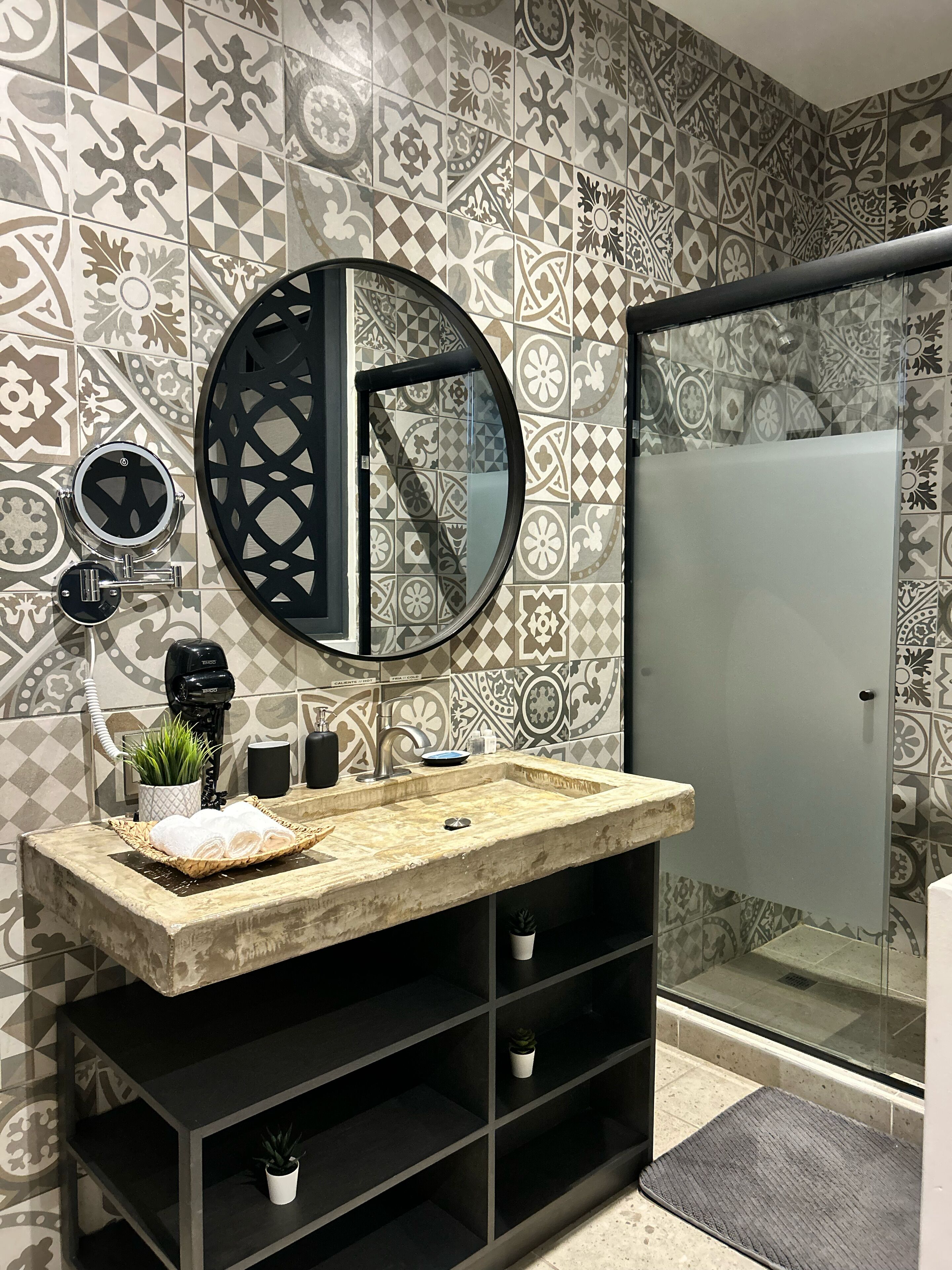 A bathroom with patterned tiles, a round mirror, a sink with a marble countertop, and a black shelf unit.