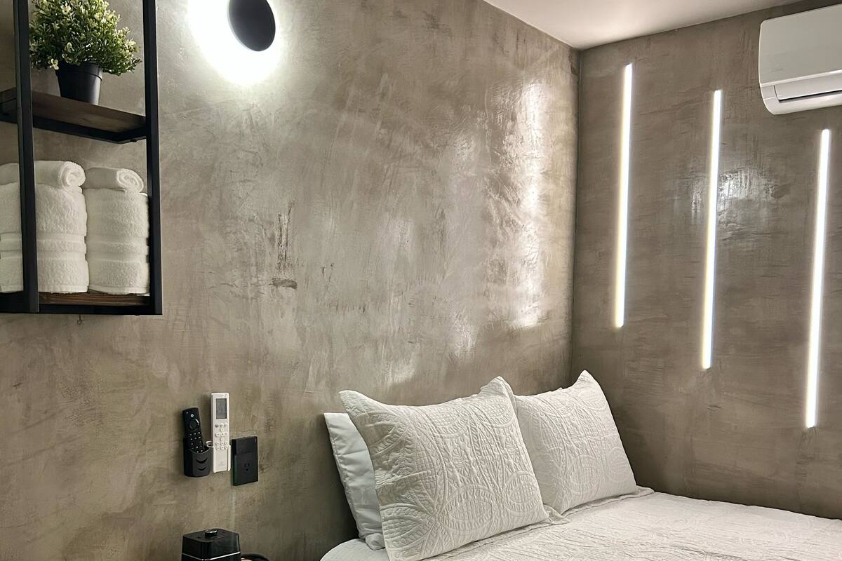 Um quarto com cama, mesa de cabeceira com abajur, prateleira na parede com toalhas e ar-condicionado fixado na parede.