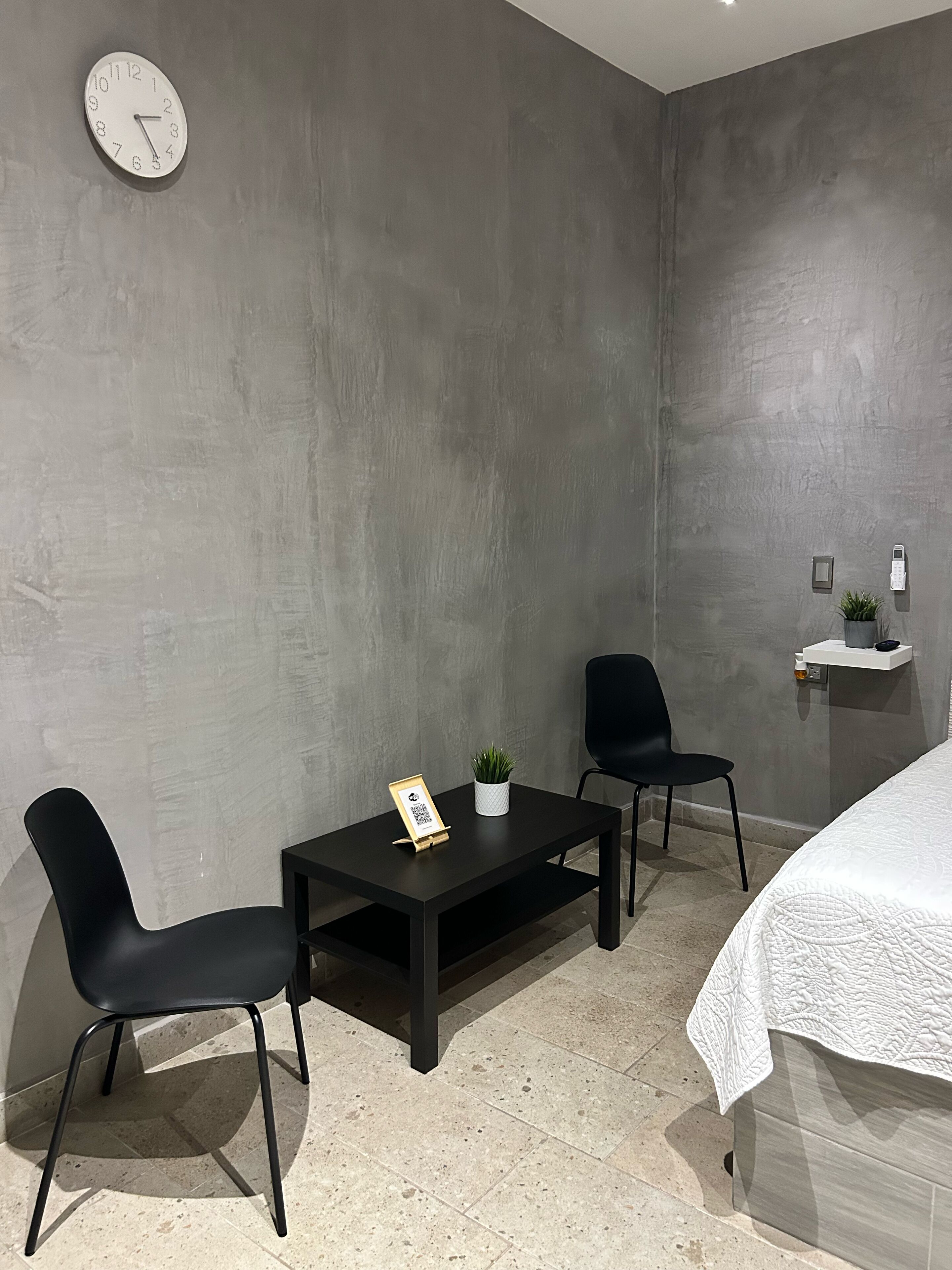 A modern hotel room with a grey concrete wall, a bed with a white quilt, two black chairs, a small table with a plant and a QR code, a wall clock, and a small shelf with a phone and a plant.