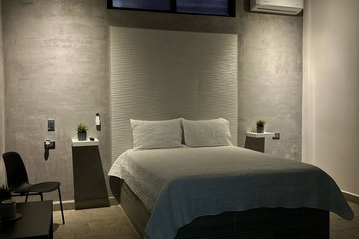Quarto moderno com cama grande, escrivaninha e cadeira. Há uma pequena mesa com uma planta e ar condicionado fixado na parede.