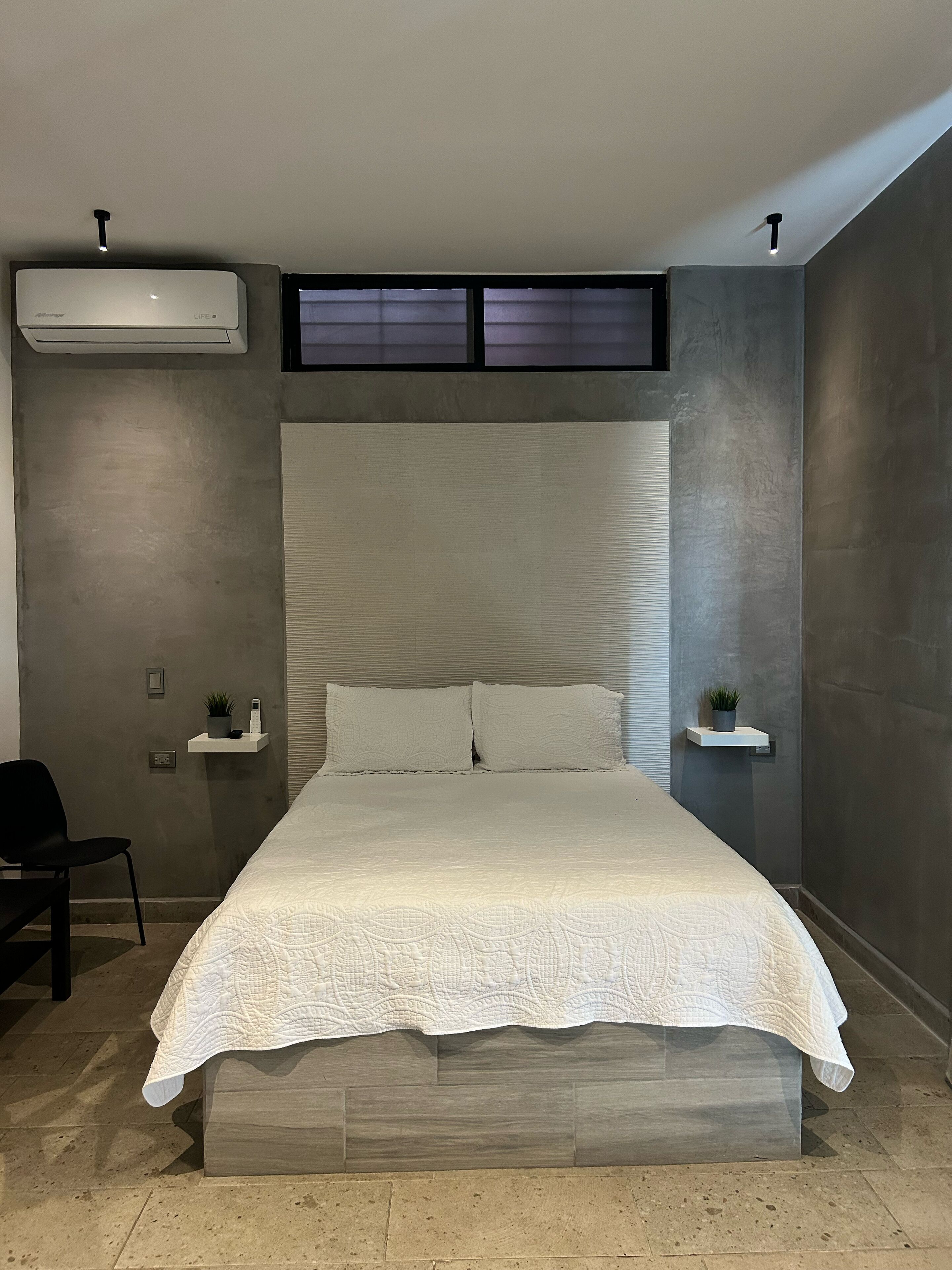 Un dormitorio moderno con una cama grande, dos mesitas de noche con plantas, un aire acondicionado de pared y una lámpara de techo.