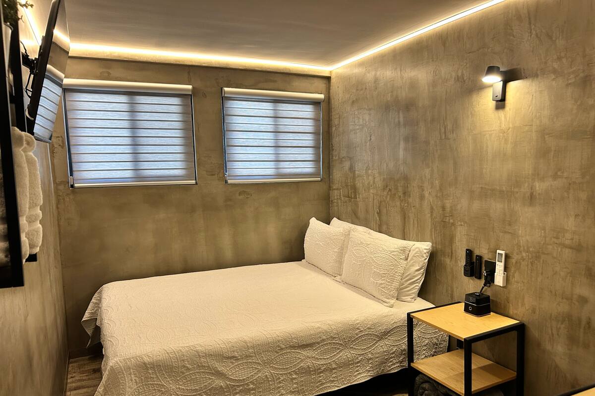 Quarto com cama, duas janelas com persianas, mesa de cabeceira com telefone e luminária na parede.
