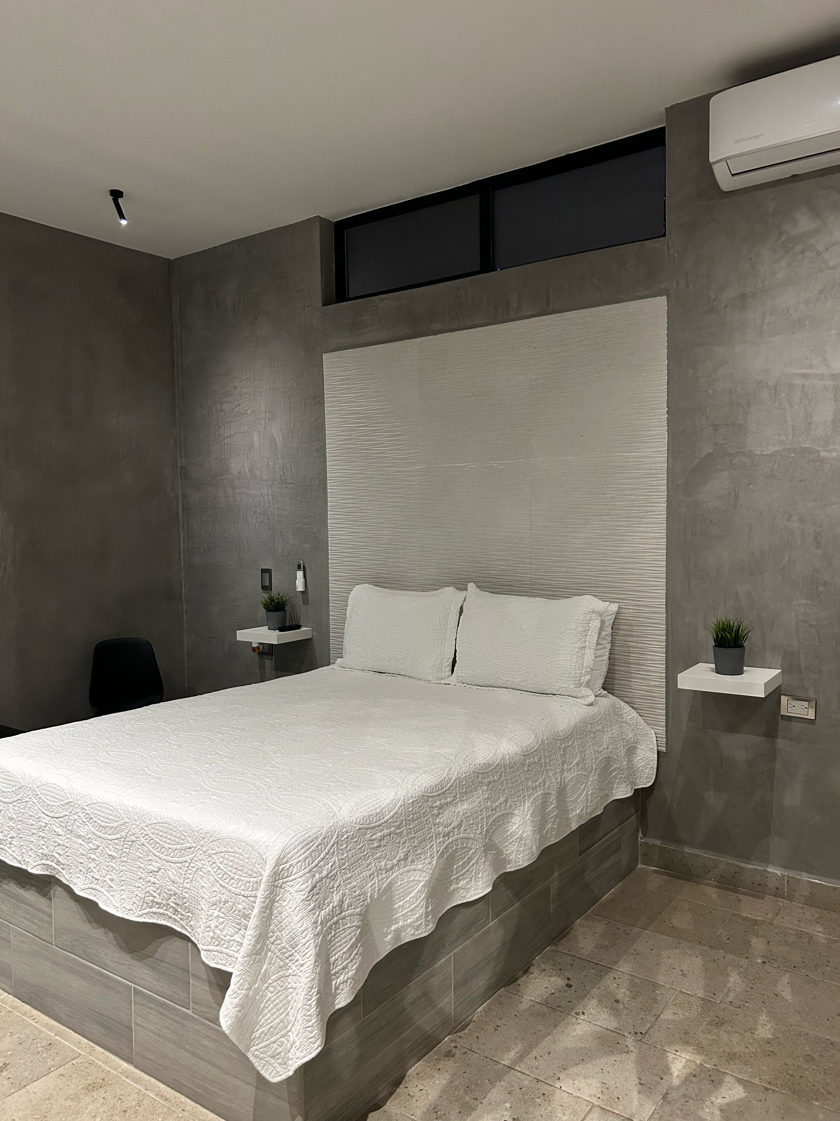 A modern bedroom with a large bed, a small bedside table, a wall-mounted air conditioner, and a potted plant.