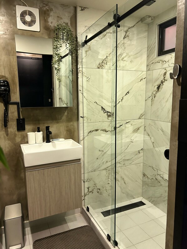 A modern bathroom with a glass shower enclosure, a marble-tiled wall, a sink with toiletries, and a hairdryer on the wall.