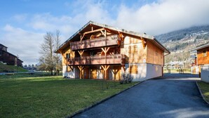 A wooden chalet with a balcony, a paved driveway, and a mountainous backdrop.