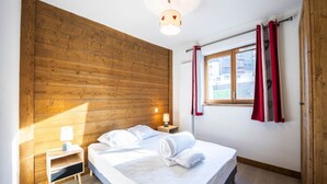 A bedroom with a wooden headboard, a bed with white linens, a nightstand with a lamp, and a window with red curtains.