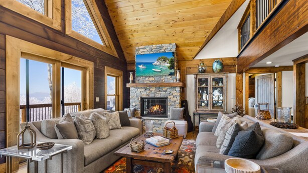 A cozy living room with a stone fireplace, large windows, and a high wooden ceiling. - 5BR Lodge w/ Hot Tub, Game Room, Great Views & Pet-Friendly (Banner Elk)