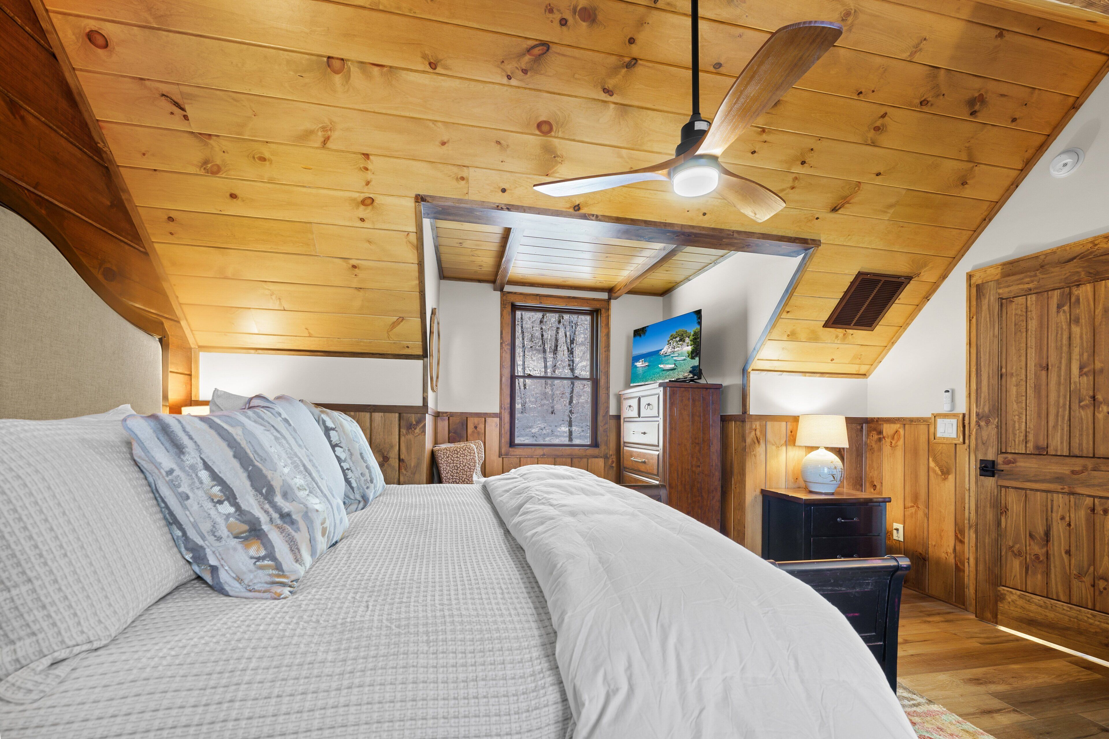 A bedroom with a wooden ceiling, a bed with white bedding, a ceiling fan, a window, and a television.