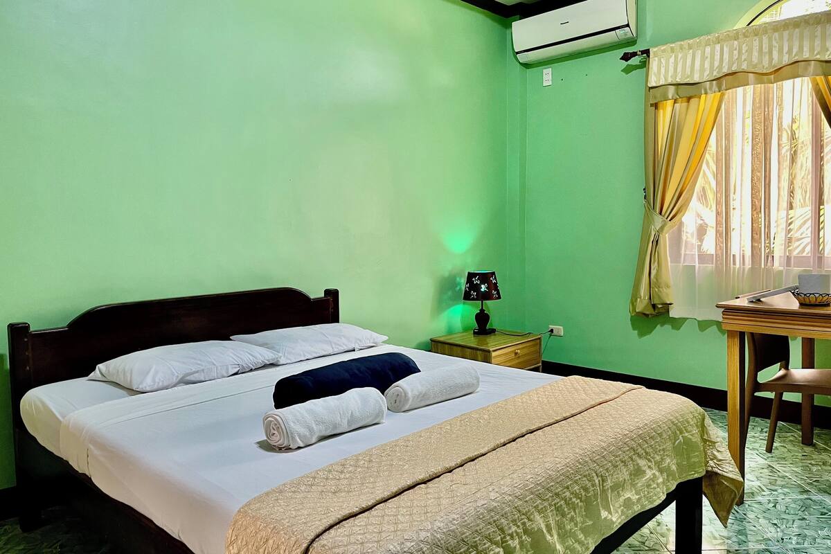 A neatly made bed with white linens and a beige quilt, a bedside lamp, and a small wooden nightstand with a lamp.