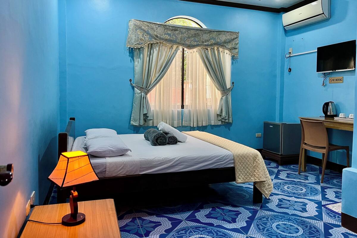 A hotel room with a bed, a desk, a chair, a lamp, and a TV.