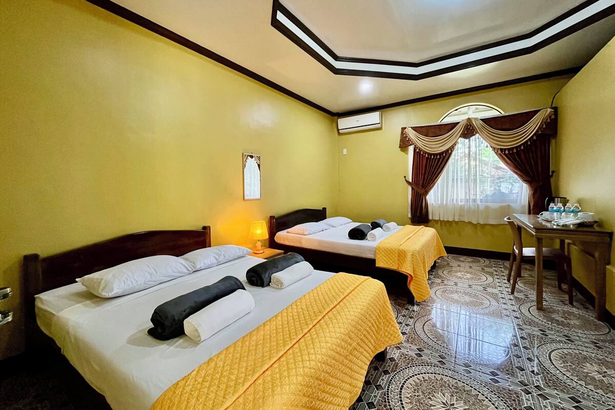 A hotel room with two beds, a desk, a chair, a window with curtains, and a patterned floor.