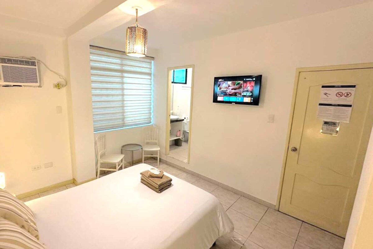 A hotel room with a bed, a TV, a window with blinds, a mirror, a door, and an air conditioning unit.