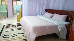 Un dormitorio con cama, cabecero, mesita de noche y cortinas en la ventana. Se ve un baño a través de una puerta abierta.
