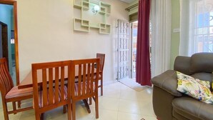 Una sala de estar con un sofá de cuero marrón, una mesa de comedor de madera con sillas, un reloj en la pared y una puerta corrediza de vidrio con cortinas.