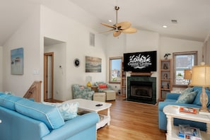 A spacious living room with a fireplace, a ceiling fan, and a large TV screen displaying 'Coastal Carolina Vacations and Sales'. - A Bbitt Beachy: Oceanside, Hot Tub, Community Pool, Easy Beach Access (Nags Head)