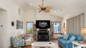 A living room with a fireplace, blue sofas, a ceiling fan, and a TV displaying 'Coastal Carolina Vacationers and Sales'. - A Bbitt Beachy: Oceanside, Hot Tub, Community Pool, Easy Beach Access (Nags Head)