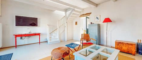 A spacious living room with a staircase, a television, a red desk, wicker chairs, a glass-top coffee table, and a red floor lamp.