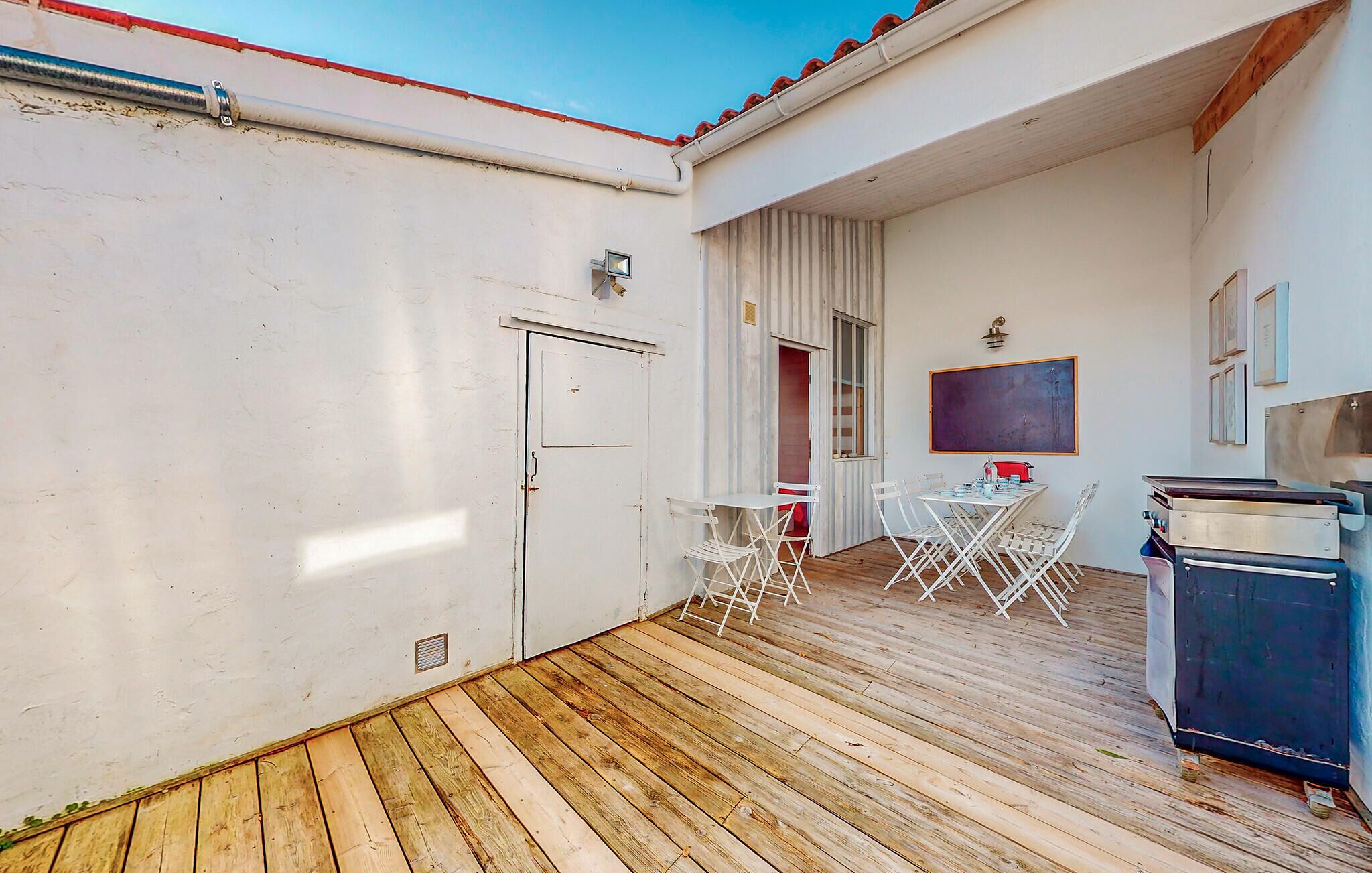 A small, enclosed patio area with a wooden floor, a white door, a small table with chairs, and a chalkboard on the wall.