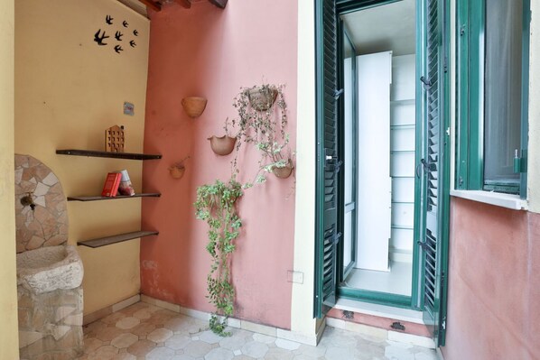Rincón de una habitación con pared rosa, un lavabo de piedra, estantes con libros y una puerta con contraventanas verdes.