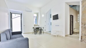 A modern living room with a grey sofa, a dining area with a table and chairs, a TV mounted on the wall, and a kitchen area visible through an open door.