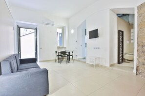 Un salon moderne avec un canapé gris, un coin repas avec une table et des chaises, un téléviseur fixé au mur et une cuisine visible à travers une porte ouverte.