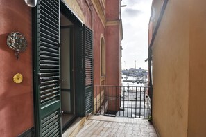 Un estrecho pasillo exterior con una reja de metal, una puerta con un cerrojo verde y una vista a la calle con edificios y un puente en la distancia.