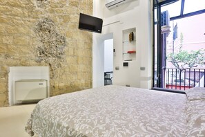 A bedroom with a stone wall, a flat-screen TV, a bed with a patterned bedspread, a white radiator, and a balcony with a railing.