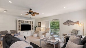 A spacious living room with a ceiling fan, a flat-screen TV, and a large fish decoration on the wall. There are multiple seating areas with sofas and chairs, and a small table with books and a lamp. - Pool View! Dog-friendly | Easy Walk to Beach, Restaurants, Village, and Pier. (Saint Simons Island)