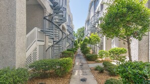 A narrow pathway flanked by multi-story buildings with external staircases, surrounded by greenery and trees. - Dog Friendly, Steps away from the beach, Restaurants, Village, and Pier. (Saint Simons Island)