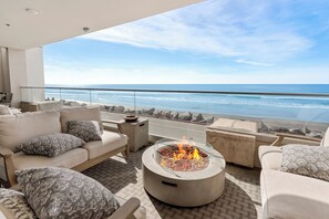 A modern outdoor seating area with a fire pit, overlooking a beach with waves and a clear sky. - The Penthouse Oceanfront (Oceanside)