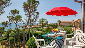 A balcony with a table set for dining, overlooking a coastal area with trees and houses. - Boudi..ou by Interhome (Anthéor)