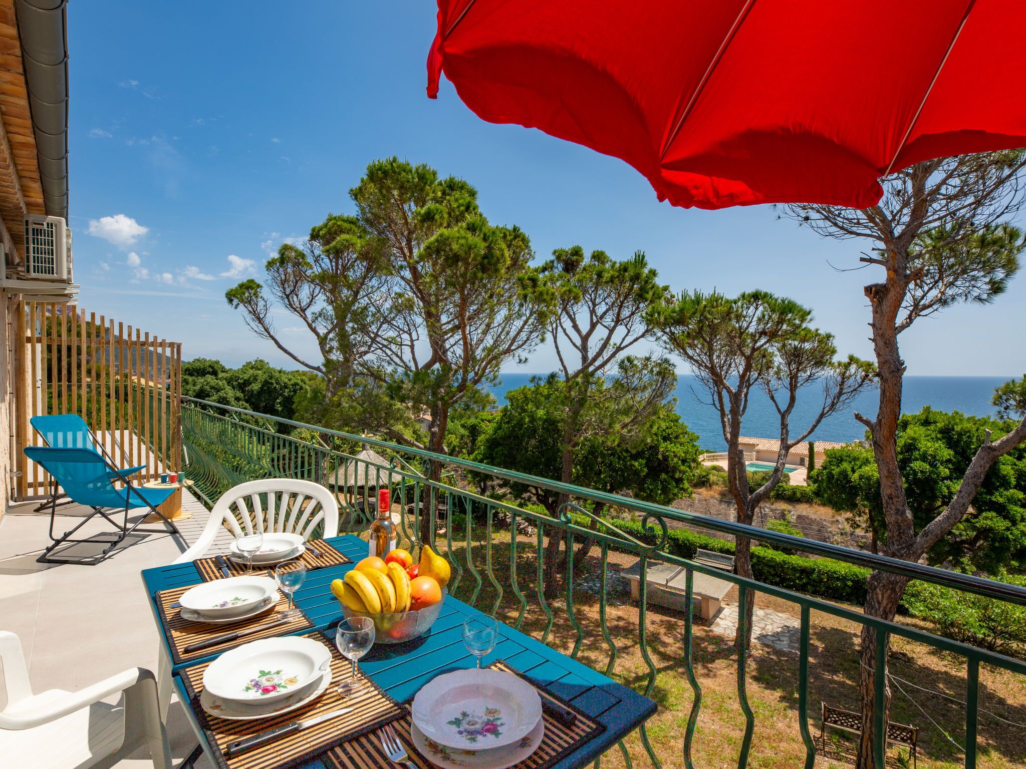 A table set outdoors with a view of the sea, a red umbrella, and a variety of fruits.