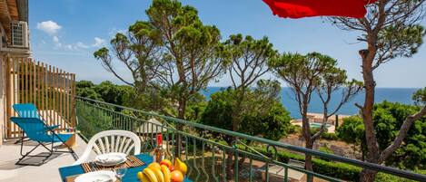 Mesa posta ao ar livre com vista para o mar, um guarda-sol vermelho e uma variedade de frutas.
