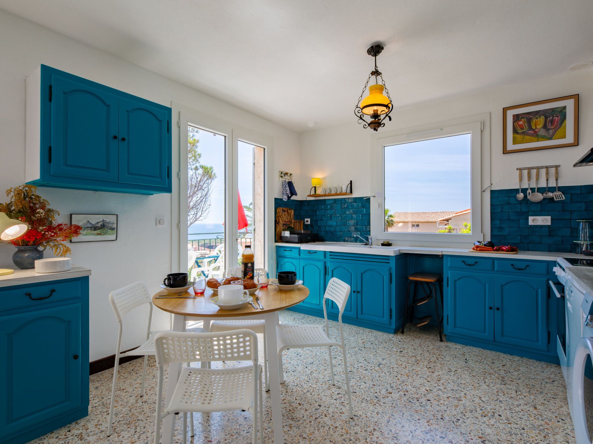 A kitchen with blue cabinets, a white dining table with chairs, a hanging light fixture, and a view through the window.