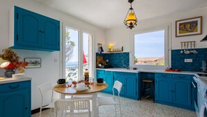 A kitchen with blue cabinets, a white dining table with chairs, a hanging light fixture, and a view through the window. - Boudi..ou by Interhome (Anthéor)