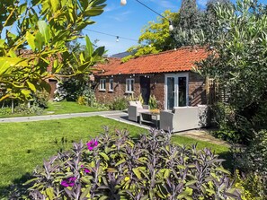 Een huis met een rood pannendak, een terras met tuinmeubilair en een verzorgde tuin met paarse bloemen.