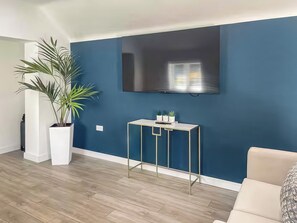A modern living room with a large flat-screen TV mounted on a dark blue wall, a white console table with decorative items, a large potted plant, and a beige sofa.