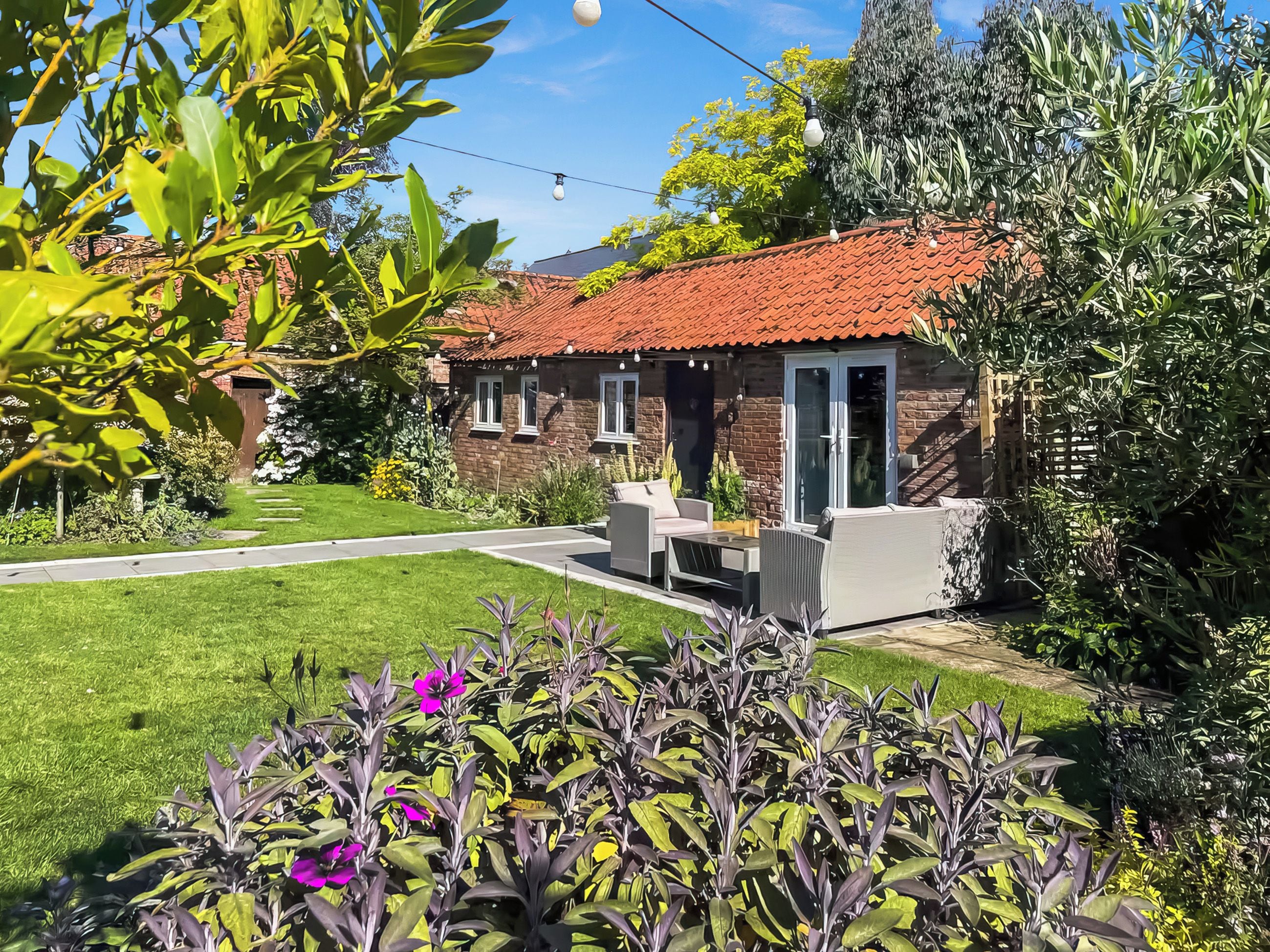 A house with a red-tiled roof, a patio with outdoor furniture, and a well-maintained garden with purple flowers.