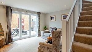 Un salon avec un escalier, un canapé, un fauteuil avec un ours en peluche, et une vue sur l’extérieur à travers une porte vitrée.