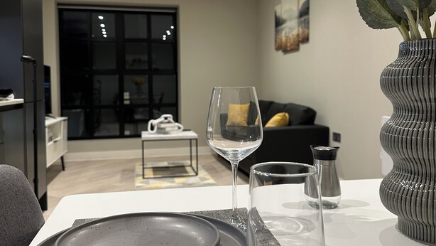 Uma área de refeições com uma mesa posta para dois, um vaso com girassóis e um interior moderno com um sofá e uma janela grande.