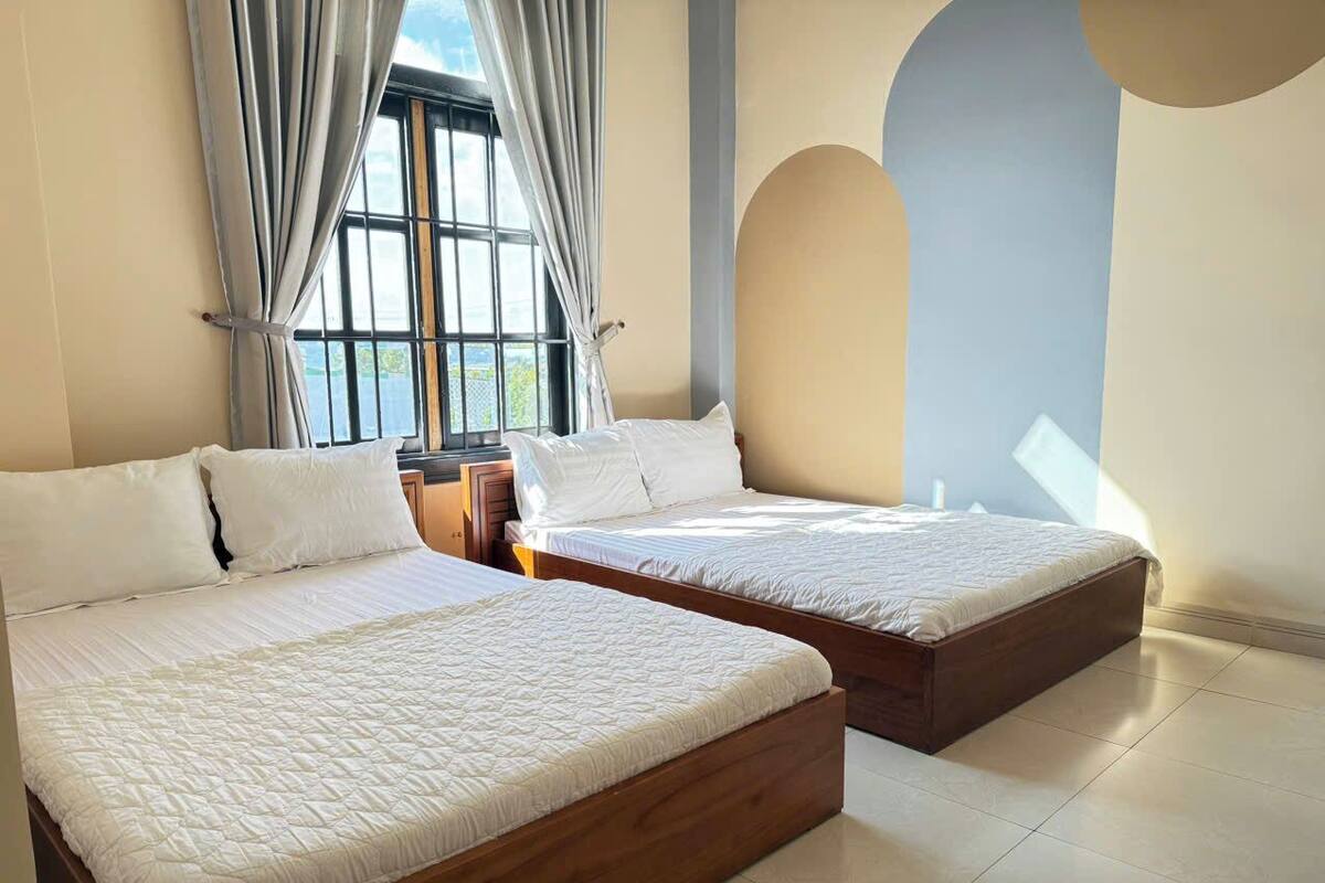 A bedroom with two wooden beds, a large window with curtains, and a view of greenery outside.